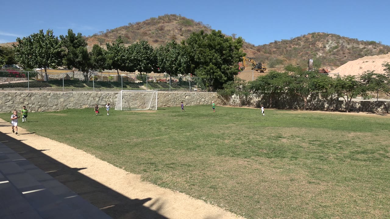 Children continue moving across the Los Cabos soccer field, with one player closer to the shaded sideline and other players near the goal.