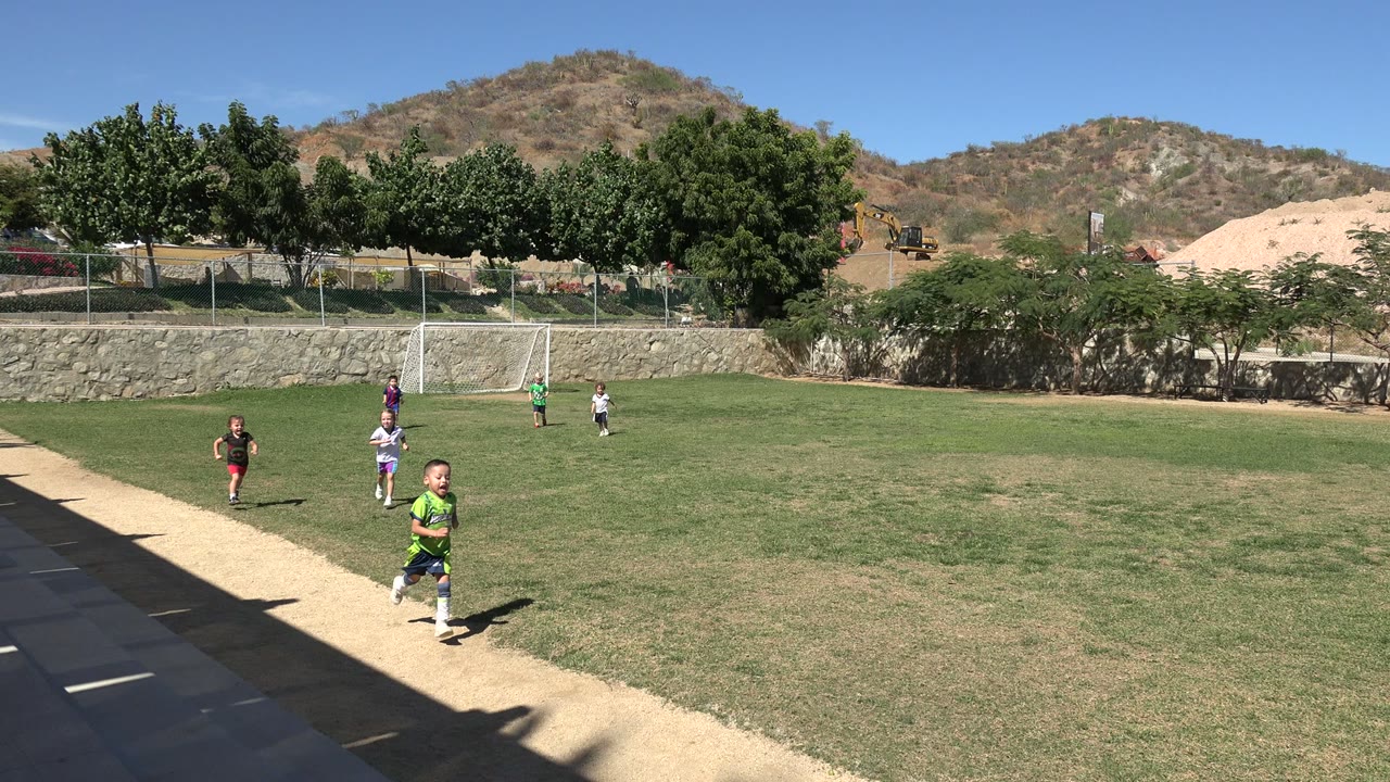 A child in a green uniform runs closest to the camera on a Los Cabos soccer field while other children move behind him near the goal.