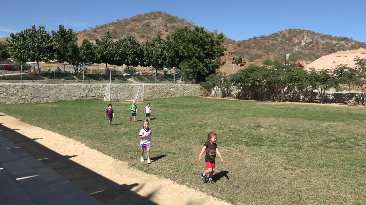 Several children stand and walk on a grass soccer field in Los Cabos, Mexico, with a small goal, trees, stone wall, and dry hills in the background.