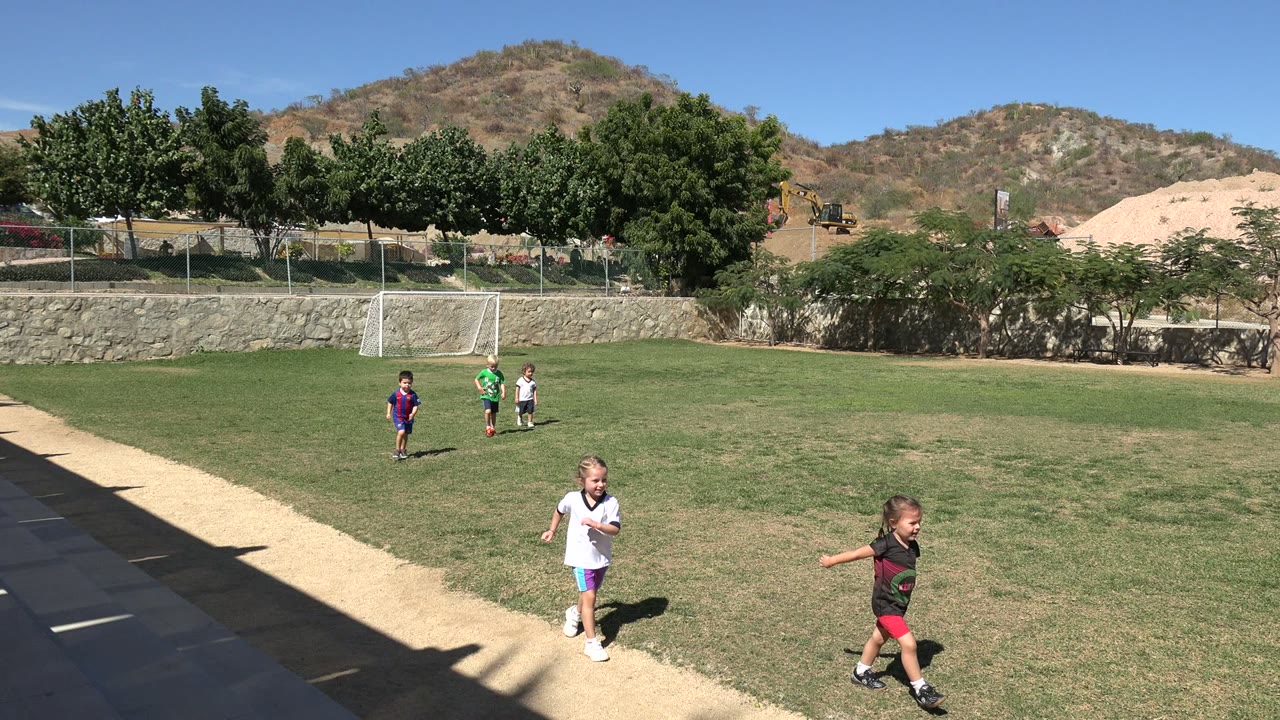 Children in colored soccer jerseys move across the Los Cabos field, with the goal near the left background and dry hills under a clear blue sky.