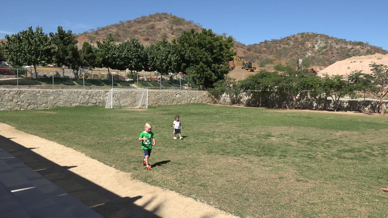 Two children on a sunny soccer field with a small goal and hills behind them.