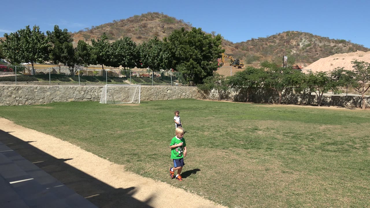 A blond child in a green jersey moves near the foreground of the Los Cabos soccer field while another child stands farther back near the grass.