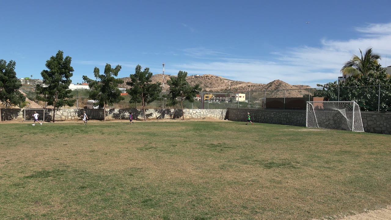 Children in light and purple sports clothing cluster near the left goal on a Los Cabos park field, with open grass, trees, stone walls, and hills beyond.