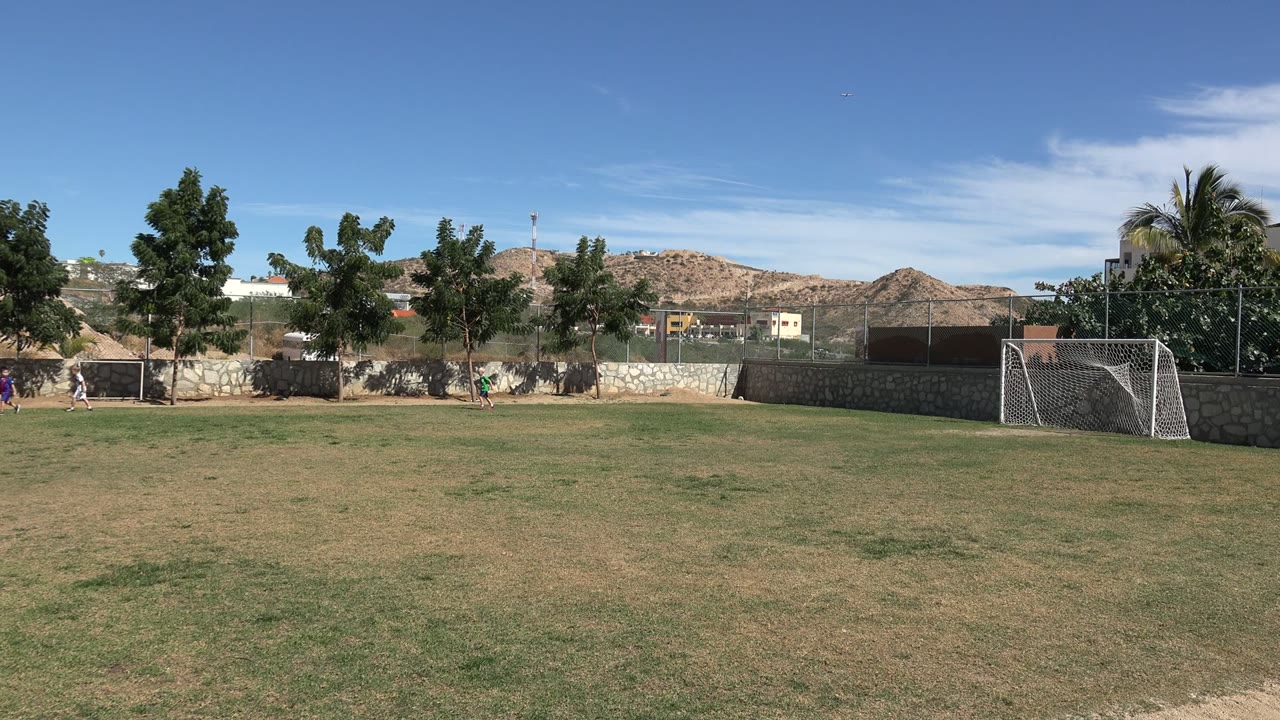 Wide view of a Los Cabos soccer field at 0:00, with open grass in front, a small goal and distant people near the left edge, a white goal at right, trees, walls, hills, and blue sky.