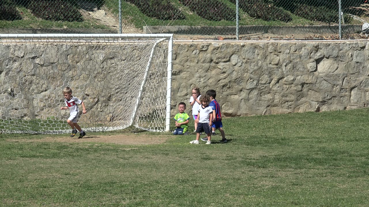 Young children gather beside the goal in Los Cabos, Mexico, while a child in green sits or kneels on the grass near the net.