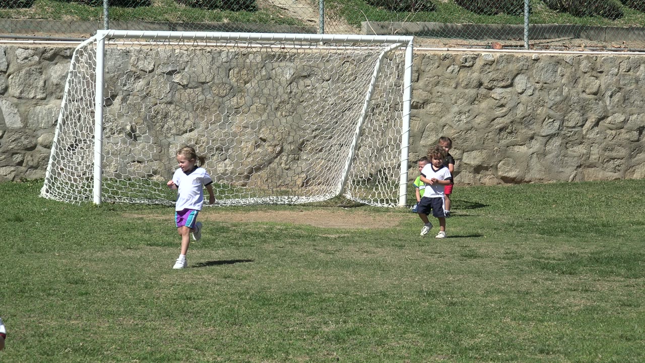 A child in a white shirt runs in front of a soccer goal in Los Cabos, Mexico, while other children stand near the net.