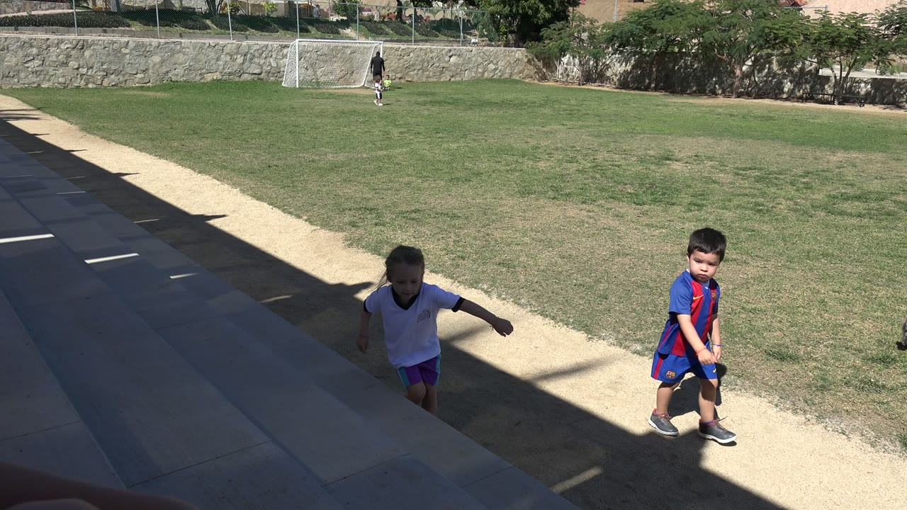 A small girl in a white shirt climbs the shaded steps while a boy in a blue and red soccer uniform walks along the field edge in Los Cabos.