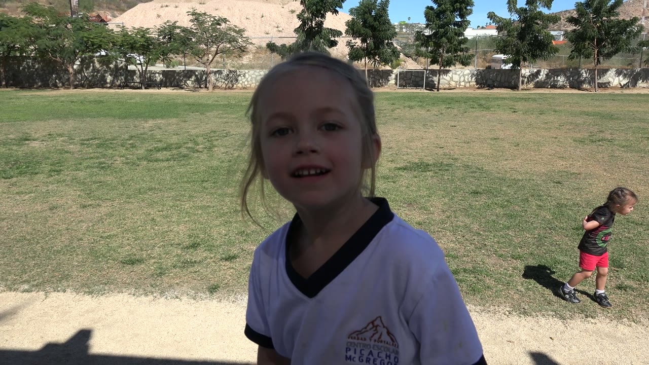 A young girl in a white shirt faces the camera from the edge of shaded steps in Los Cabos, Mexico, with patchy grass and a dirt path in the background.