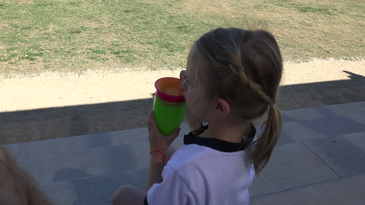 A young girl in a white shirt sits on shaded steps in Los Cabos, Mexico and drinks from a bright green cup with an orange rim as sunlit grass fills the background.