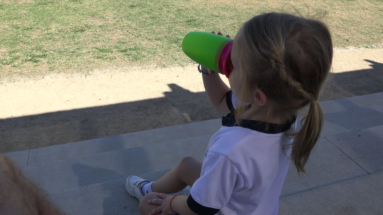 Young girl drinking from a green cup while seated on shaded pavement near grass.
