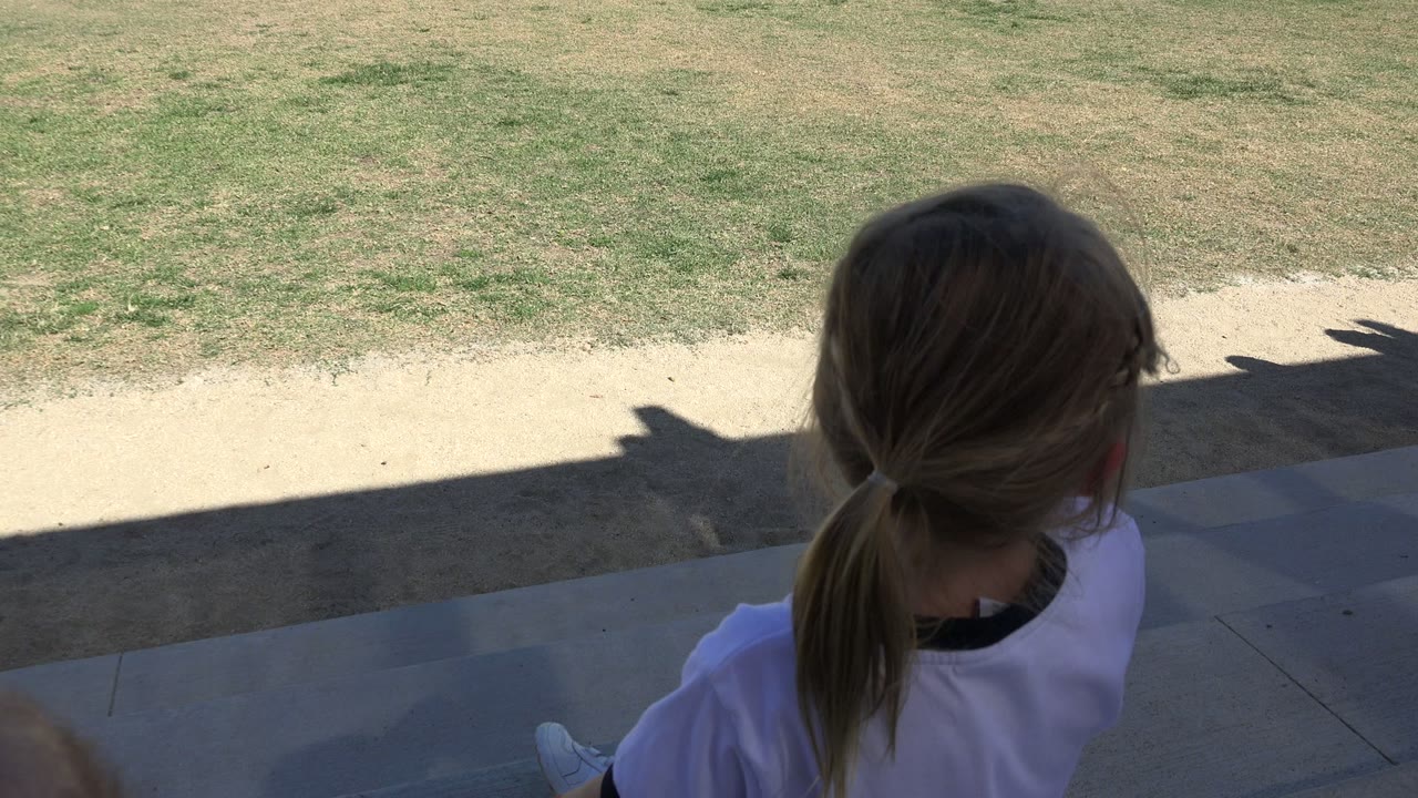 The girl in Los Cabos faces away from camera on shaded pavement, with grass and a sunlit dirt strip ahead.