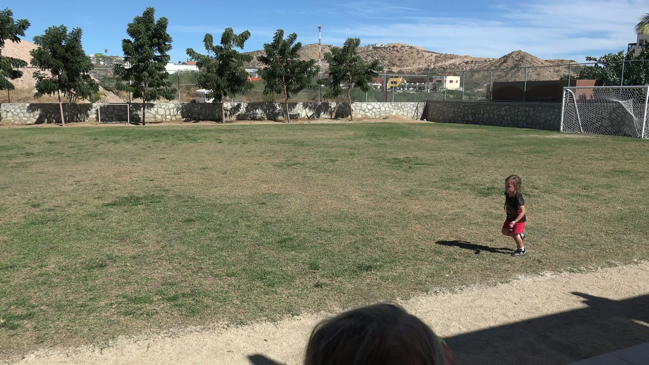 A toddler walks across the Los Cabos soccer field as another young child appears near the lower right foreground, with the goal and hillside behind them.