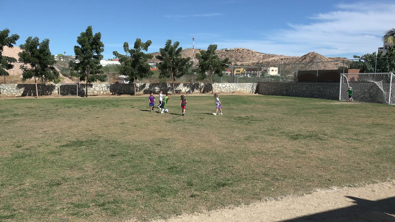 Children gather around the ball in the middle of a sunlit Los Cabos soccer field, with the white goal frame visible on the right side.