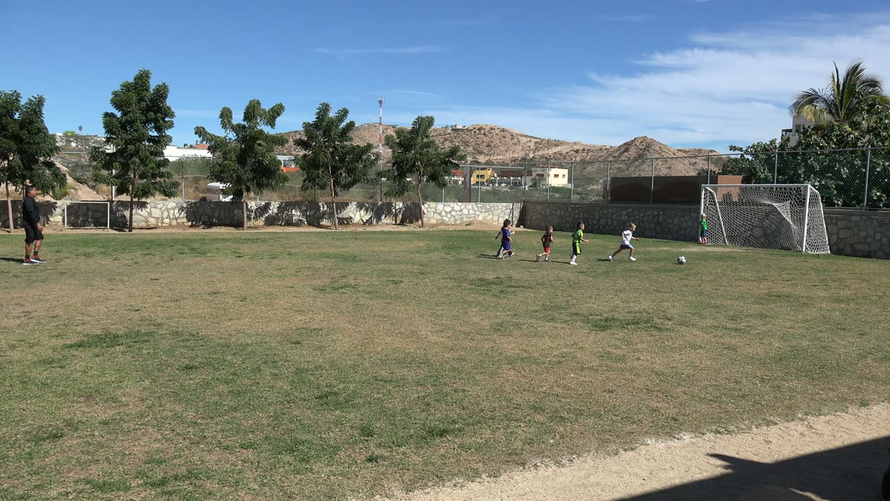 Children in several colored shirts move across the Los Cabos football field toward the goal, with patchy grass, a stone boundary wall, trees, hills, and bright blue sky visible.