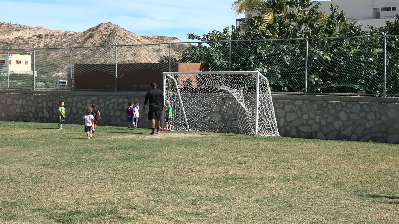 Children and a coach near a small soccer goal on a sunny grass field.