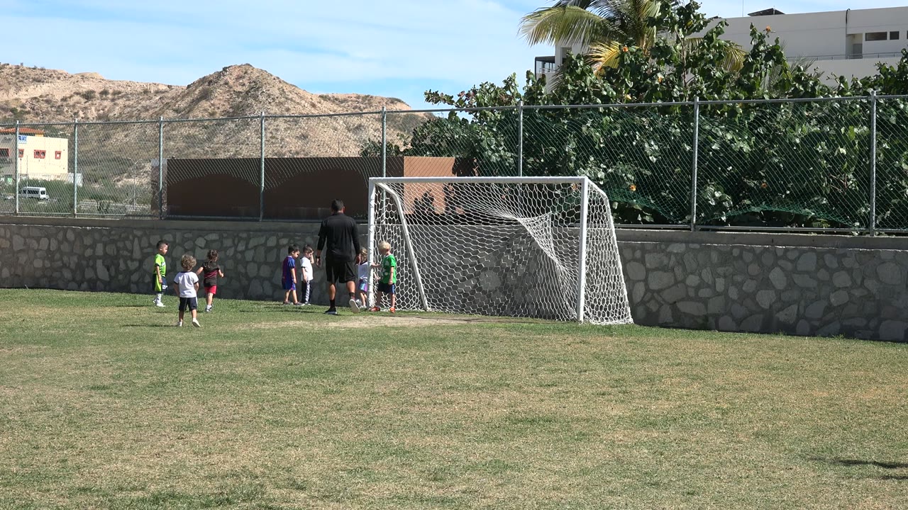 Several young soccer players cluster near the white goal in Los Cabos, Mexico, while the coach stands in dark clothing and a child in green waits near the post.