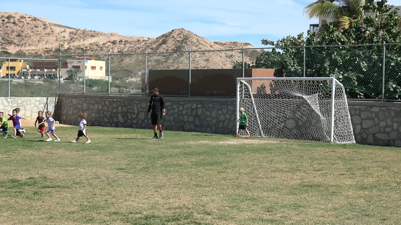 Children play soccer on a wide grass field in Los Cabos, Mexico, with a small goal, adult coach, stone wall, fence, trees, buildings, and dry hills behind them.