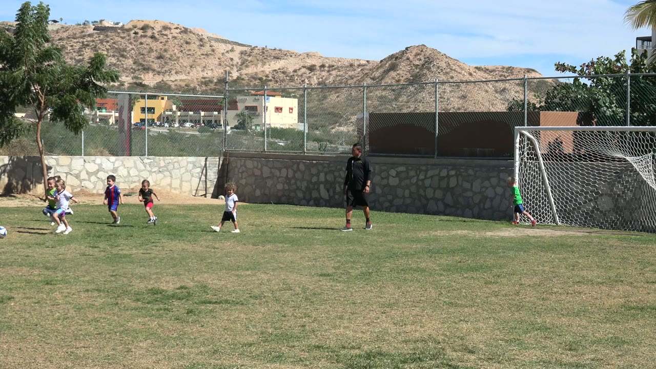 A child in white and another in dark soccer clothes run near a ball on the Los Cabos grass field while a goalkeeper stands by the net and an adult coach watches from the side.