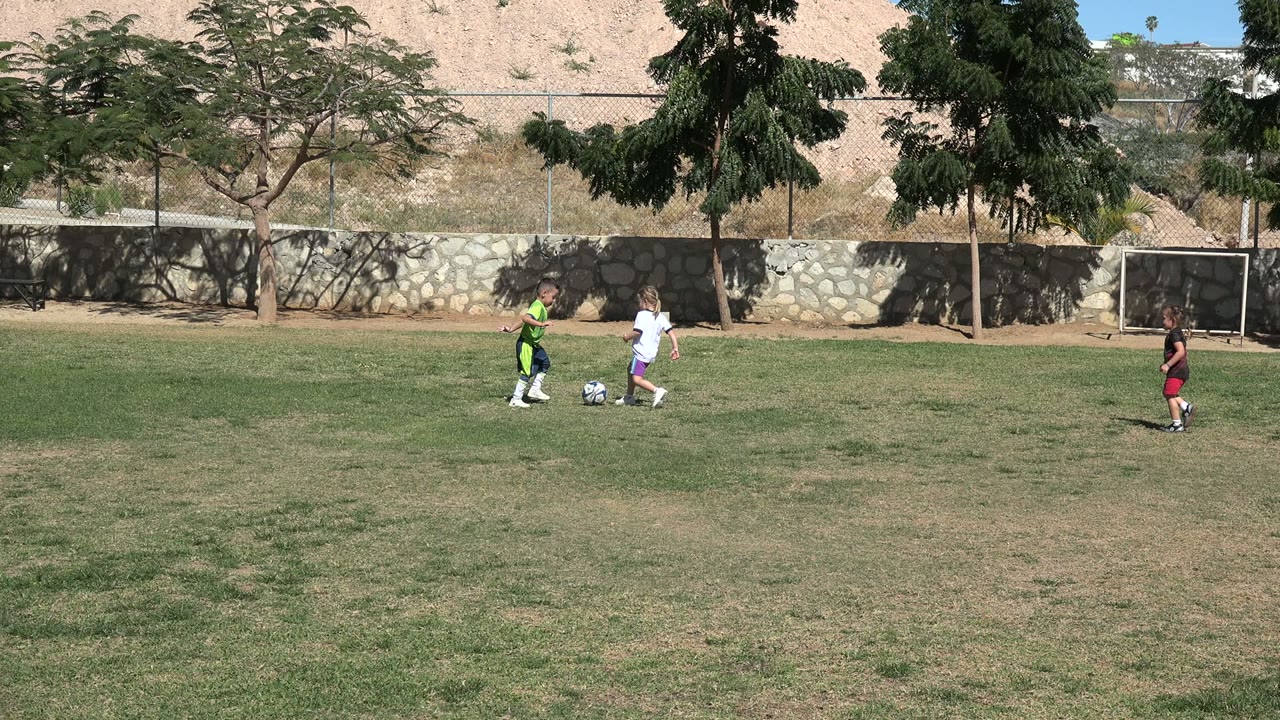 Children continue playing soccer in Los Cabos, Mexico, as the child in white runs near the ball and the boy in green approaches from the left.