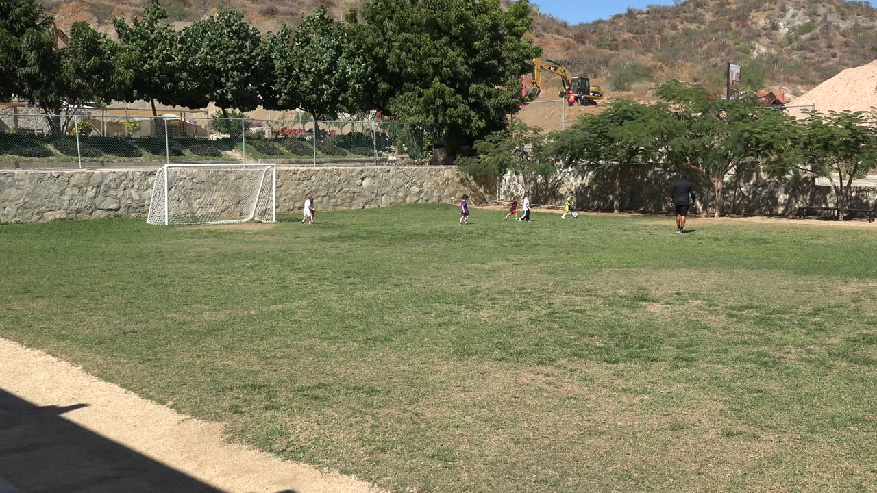 Los Cabos soccer field with a white goal left of center, children spaced across the grass, shaded trees along the back edge, and a dirt hillside beyond.
