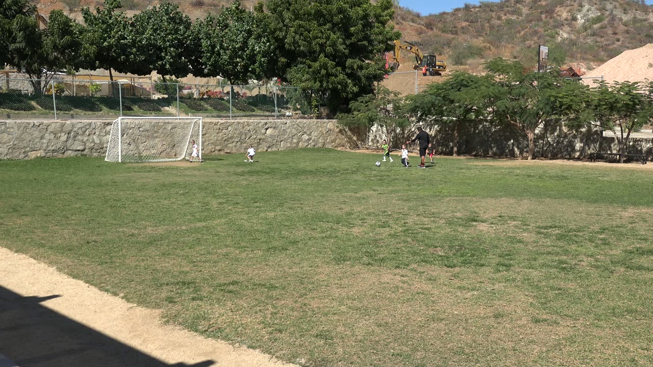 Los Cabos youth soccer practice continues with the ball in the midground, the coach standing nearby, children spaced around the play, and the white goal left of center.