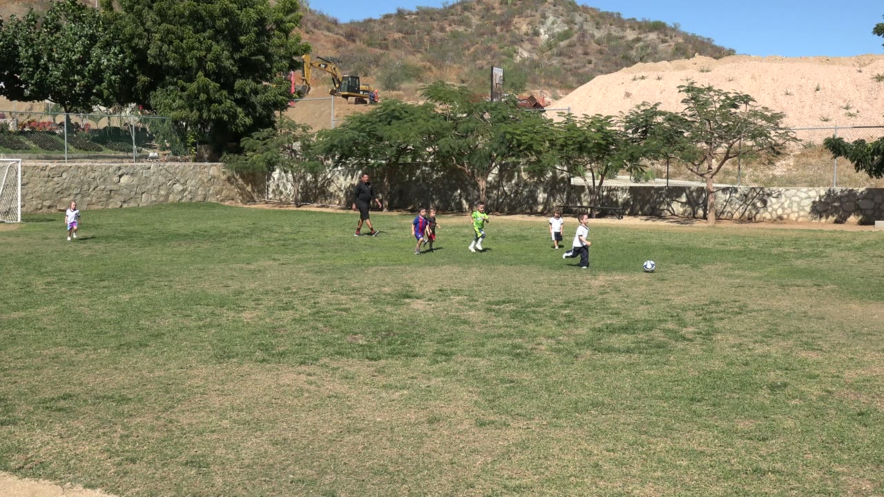 Several children in uniforms move around the soccer ball in Los Cabos, Mexico, with an adult on the field, trees and a stone wall across the background.