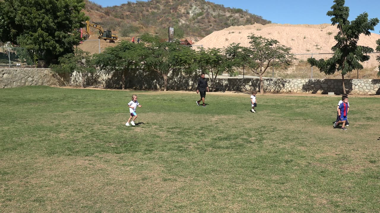 Children move across the patchy grass in Los Cabos, Mexico, with one child near center and an adult in dark clothing behind them by the wall.