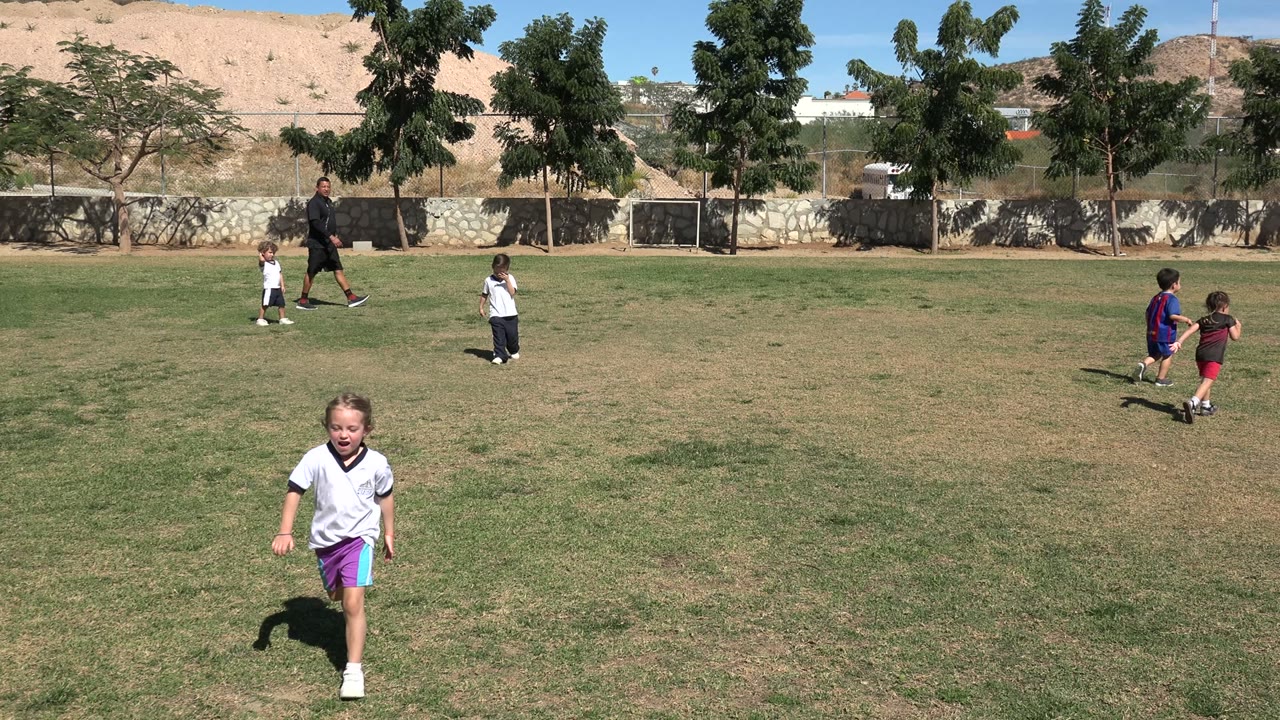 A young girl in a white shirt and purple shorts runs across a grassy field in Los Cabos, Mexico, while other children and an adult are visible near trees and a low wall.