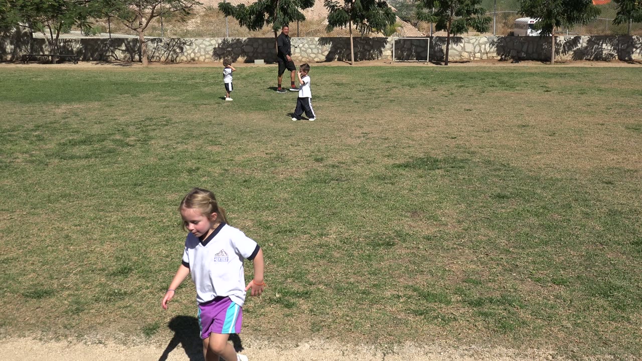The girl continues across the Los Cabos field in bright sunlight, with patchy grass, distant children, an adult, trees, and a low stone wall behind her.