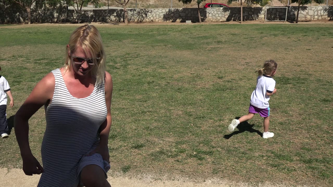 In Los Cabos, Mexico, an adult woman in sunglasses stands close to the camera while a child runs across a grass football pitch behind her.