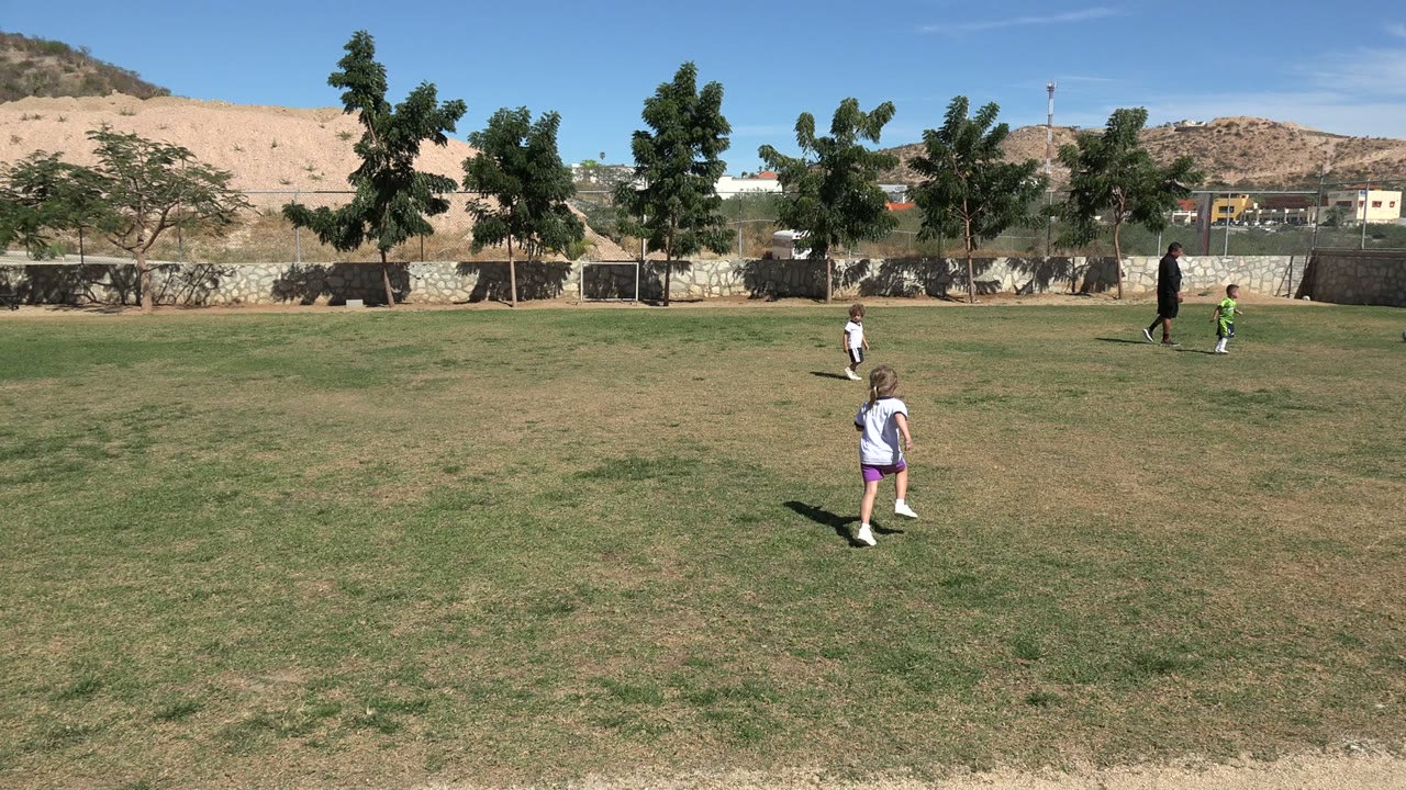 Children in white and purple sports clothes are spaced across a dry grass football field in Los Cabos, Mexico, with adults watching near the far side.