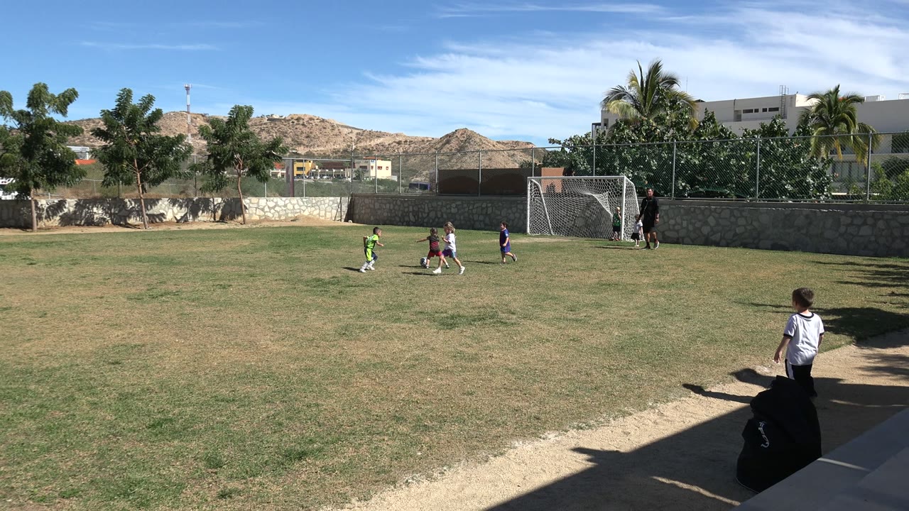 A small group of children chase a soccer ball across the grass in Los Cabos, Mexico, while a child stands near the shaded sideline and an adult remains by the goal.