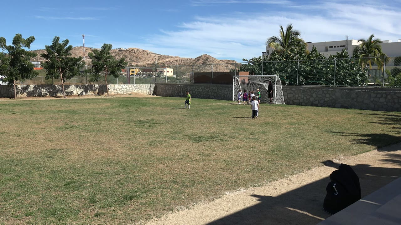 Children remain around the soccer goal and open field in Los Cabos, Mexico, with one child farther left and several players grouped by the net.