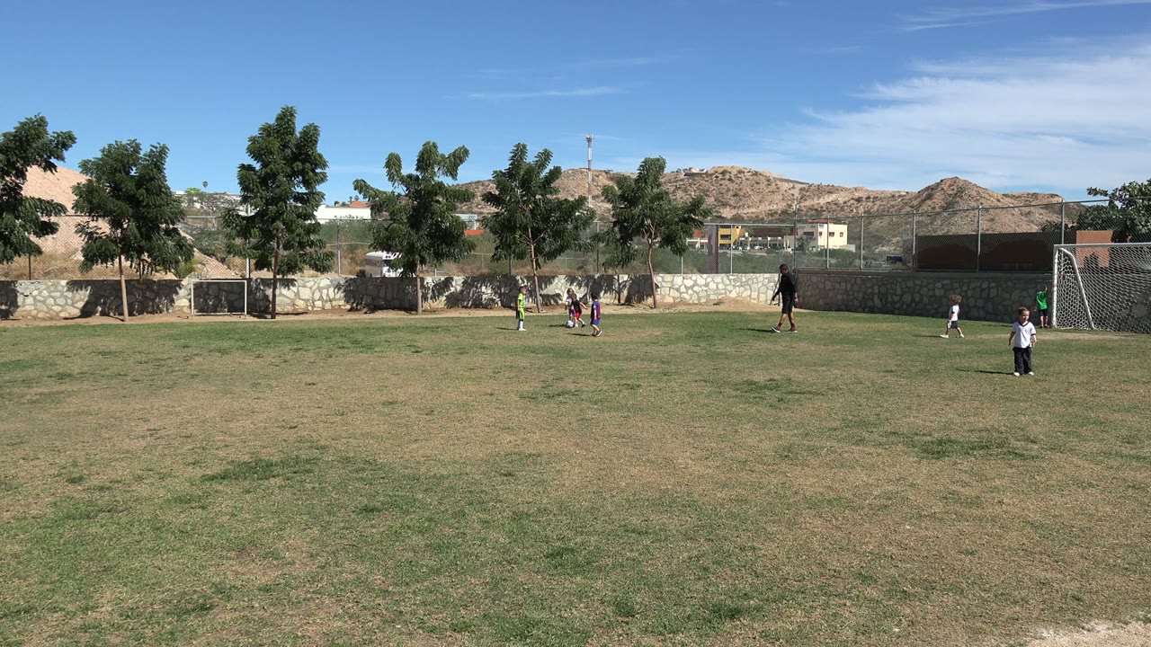 Children gather near the ball on the Los Cabos field while a coach stands to the right, with a large goal, smaller goal, trees, fencing, and hills behind them.