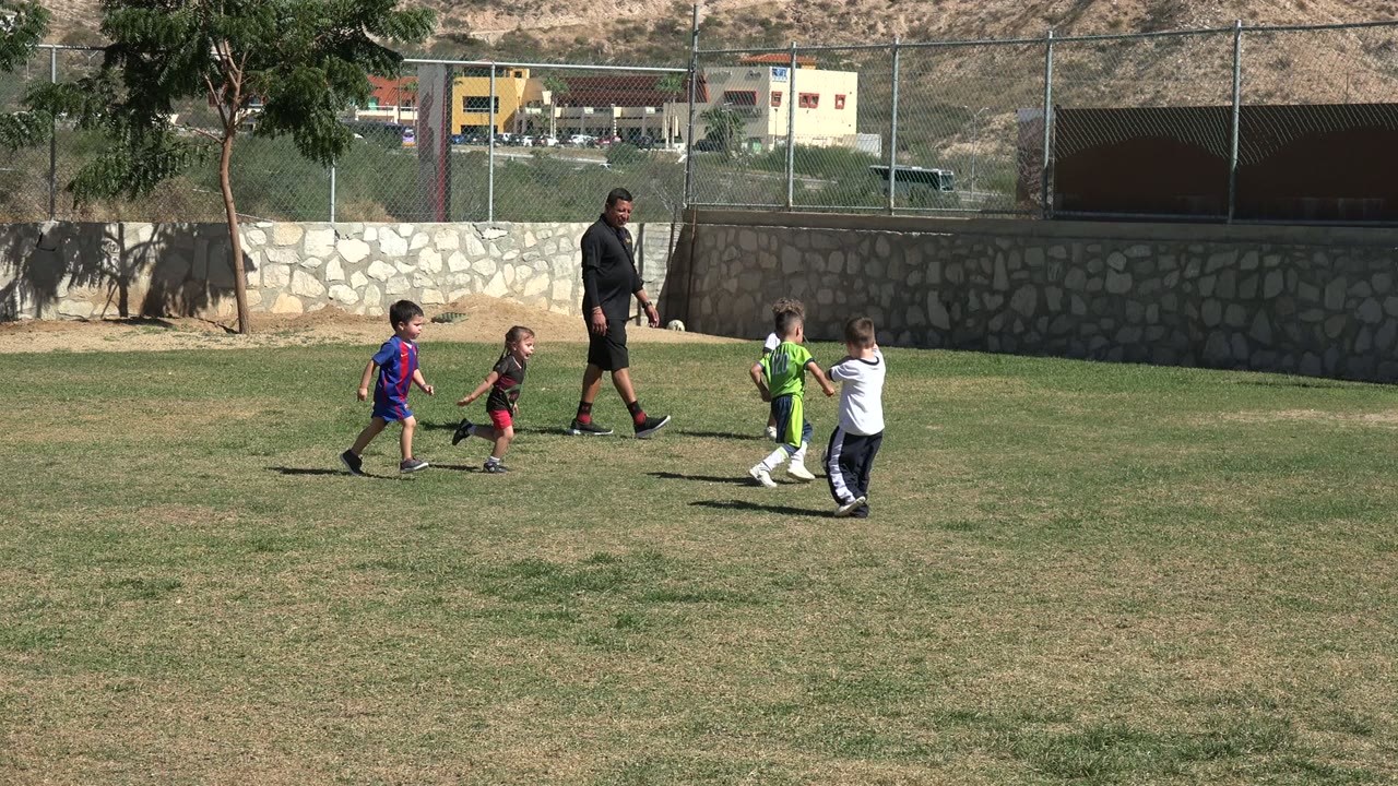 Several children chase the soccer ball across the grass in Los Cabos, Mexico, with an adult nearby and the field fence and hillside beyond.