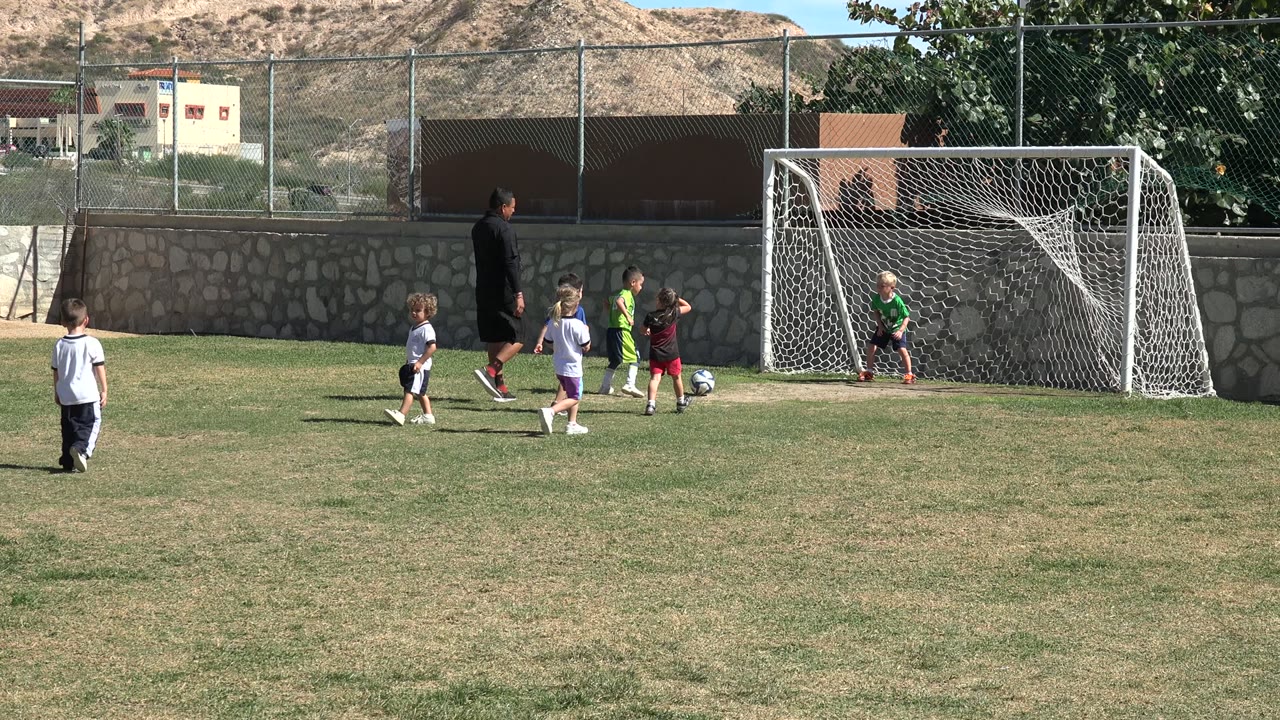 At the start of the Los Cabos clip, young children gather near a white soccer goal while an adult coach stands beside them on a sunny grass field.