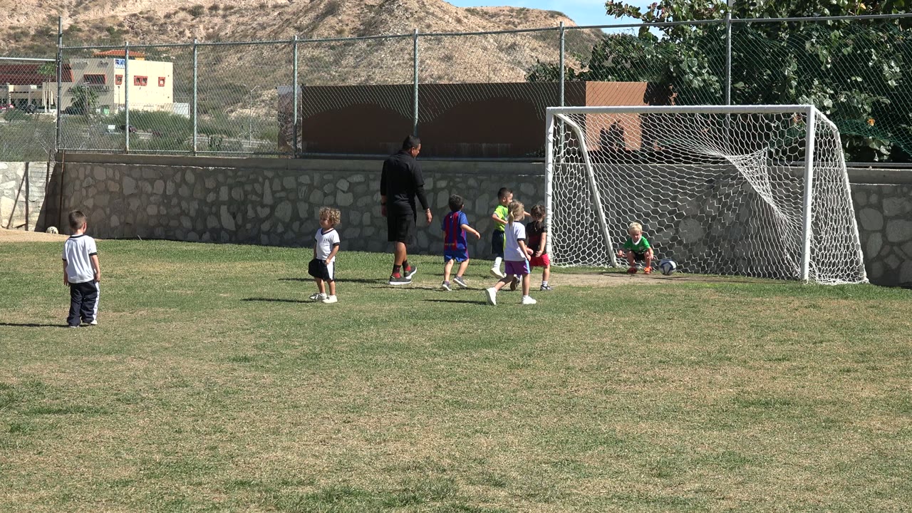 One second into the Los Cabos scene, children in colored jerseys cluster around the ball while the coach watches near the right-side goal.