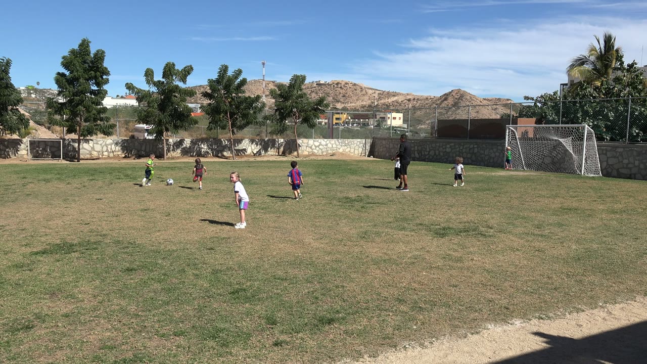 Children move around the soccer ball on a dry field in Los Cabos, Mexico, while a white goal, adult figure, trees, walls, and hillside background remain in view.