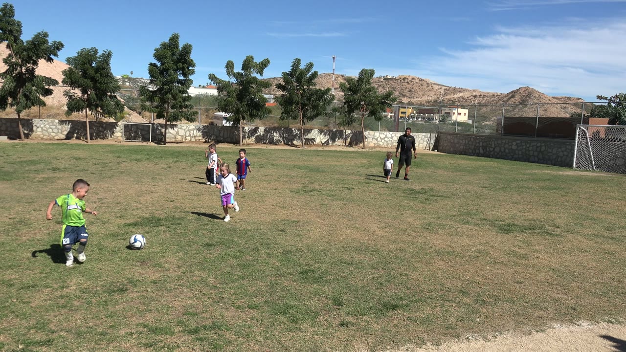 At 0:00 in Los Cabos, small children stand and move around a soccer ball on a patchy grass field, with an adult, fence, trees, buildings, dry hills, and blue sky in the background.