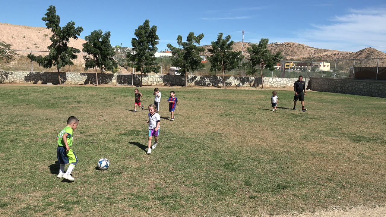 At 0:01 in Los Cabos, a child in a bright green soccer kit moves toward the ball in the foreground while several children and an adult stand across the sunny grass field.