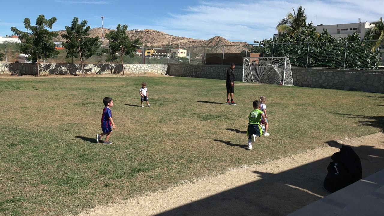 Children continue across the soccer field in Los Cabos, Mexico, with the adult coach near the goal and hills and trees behind the stone wall.