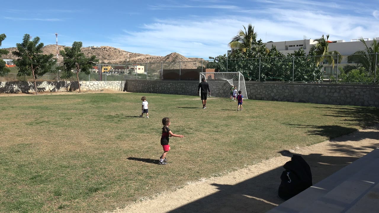 A child in red shorts crosses the foreground of a Los Cabos soccer field while other children and an adult remain near the goal under bright sun.