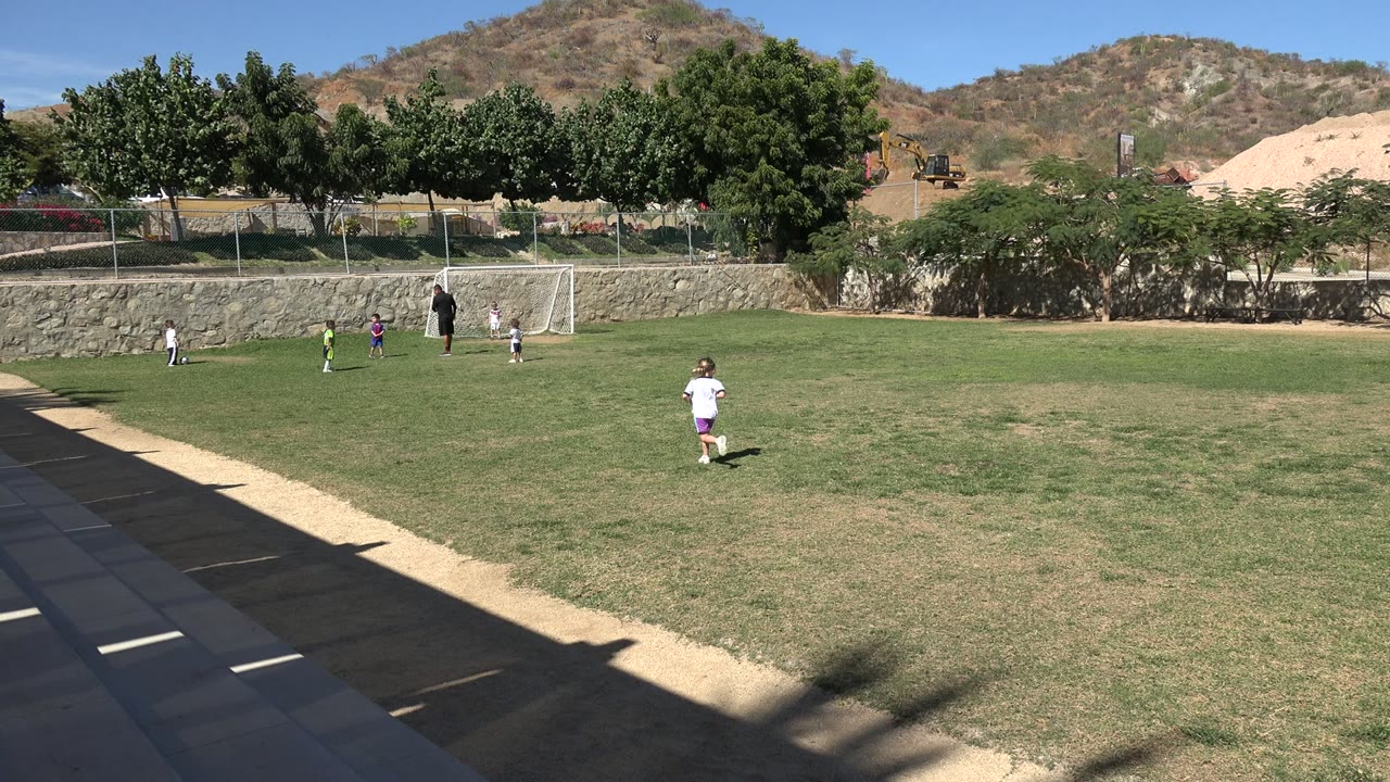 The running child has moved farther right across the Los Cabos soccer field, with several small children clustered near the goal and an adult standing beside it.