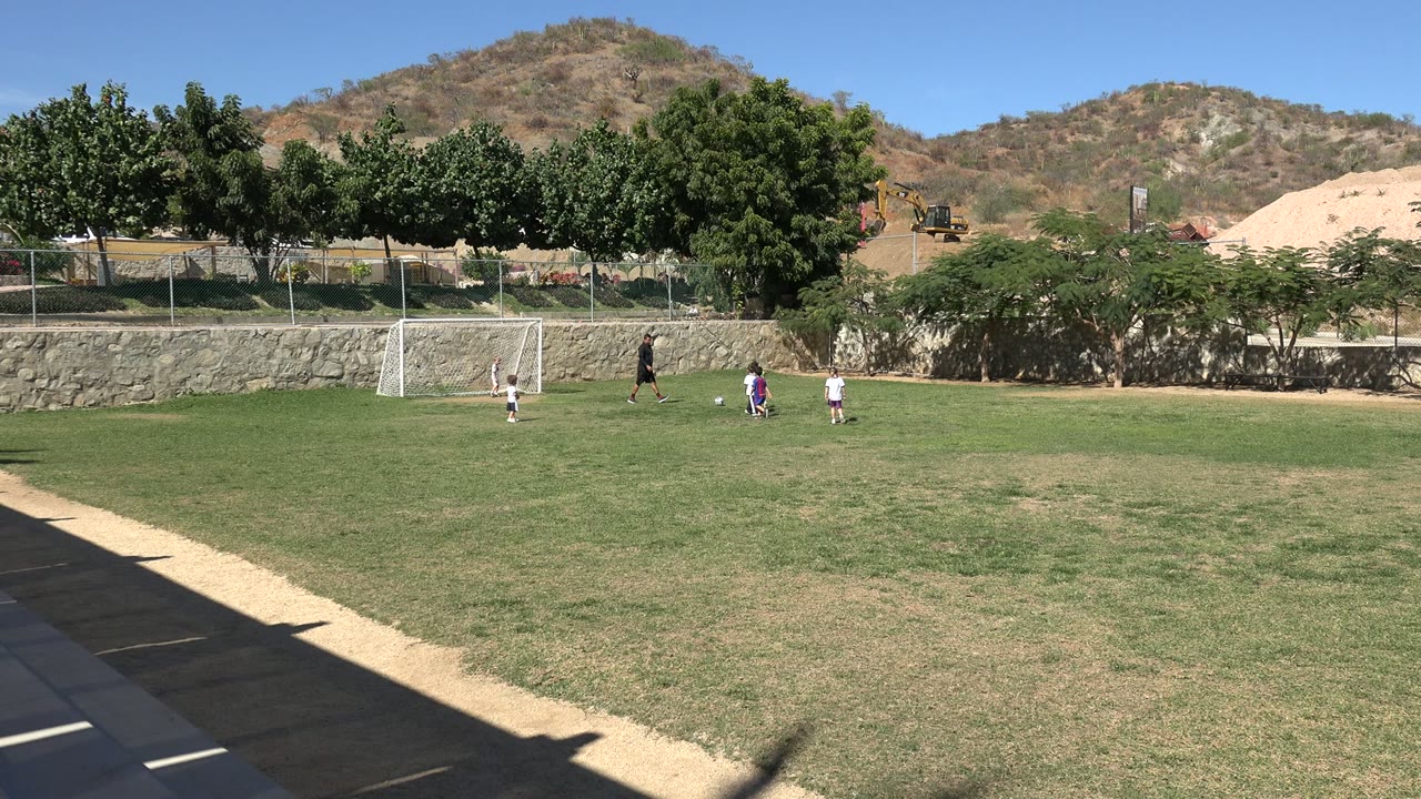 Second moment of the Los Cabos soccer clip shows the children slightly separated across the grass while the adult remains near the goal area and hills fill the background.