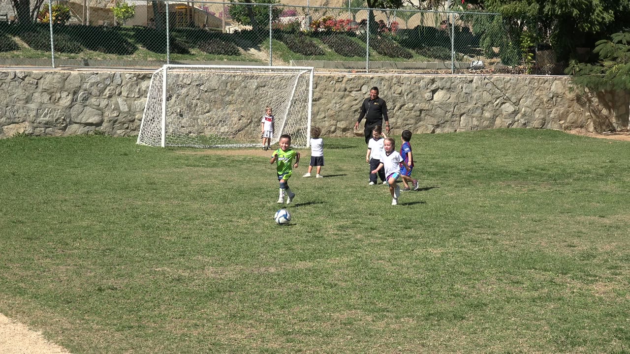 In Los Cabos, Mexico, a boy in a bright green shirt runs toward a black and white soccer ball while other children cluster near the small goal and an adult coach watches from the background.