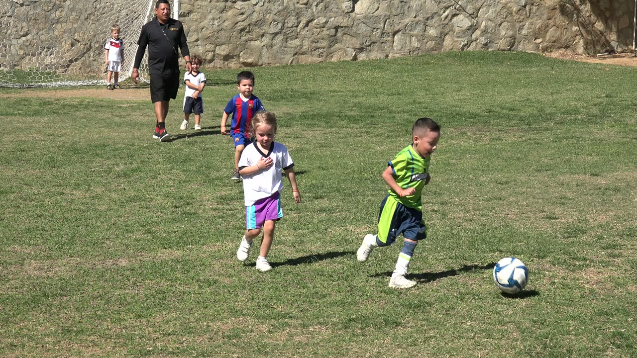 In Los Cabos, Mexico, several children and an adult are visible on a sunny grass field while a young boy in a bright green soccer uniform runs toward a blue and white ball.