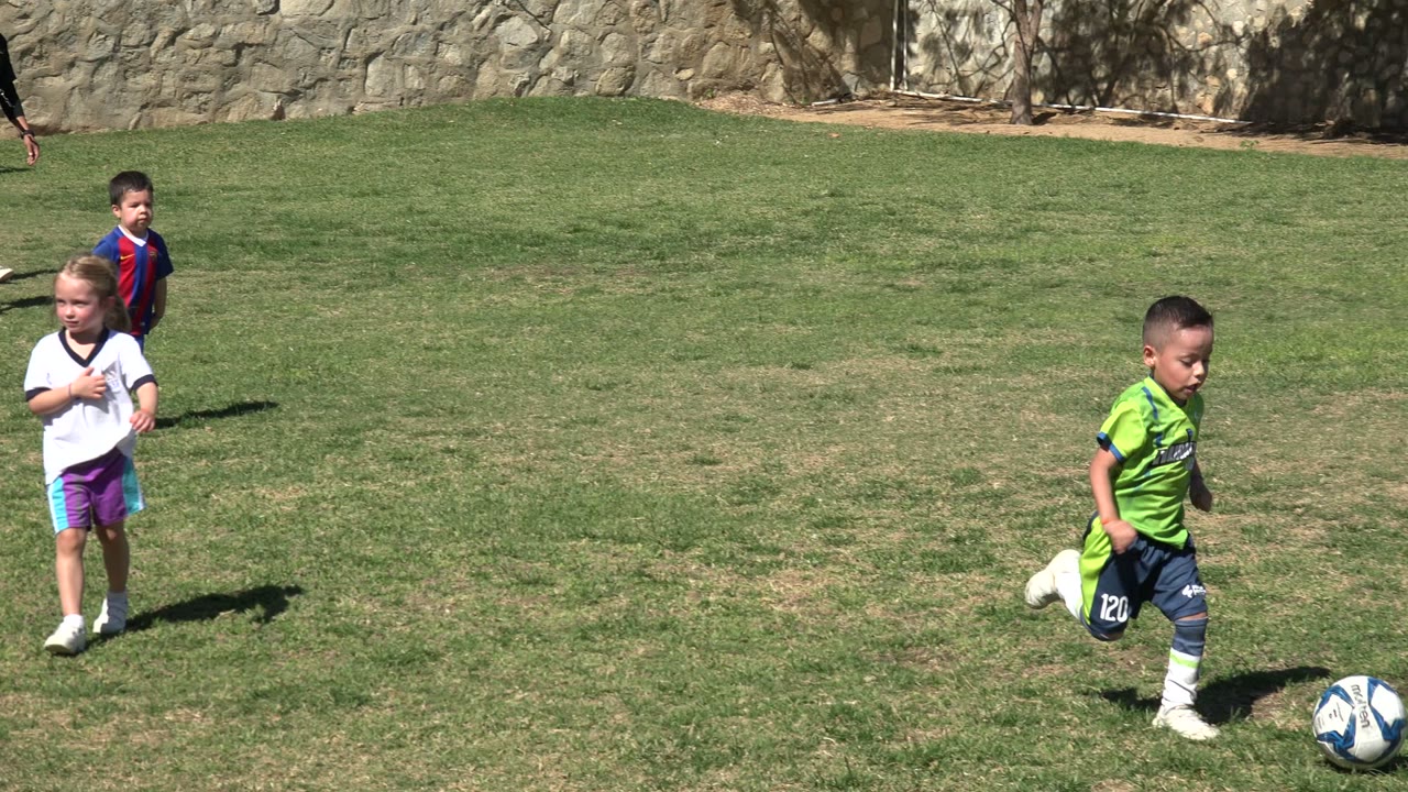 In Los Cabos, Mexico, the boy in the green uniform runs in profile across patchy grass with the soccer ball just ahead of him and trees and a stone wall behind.