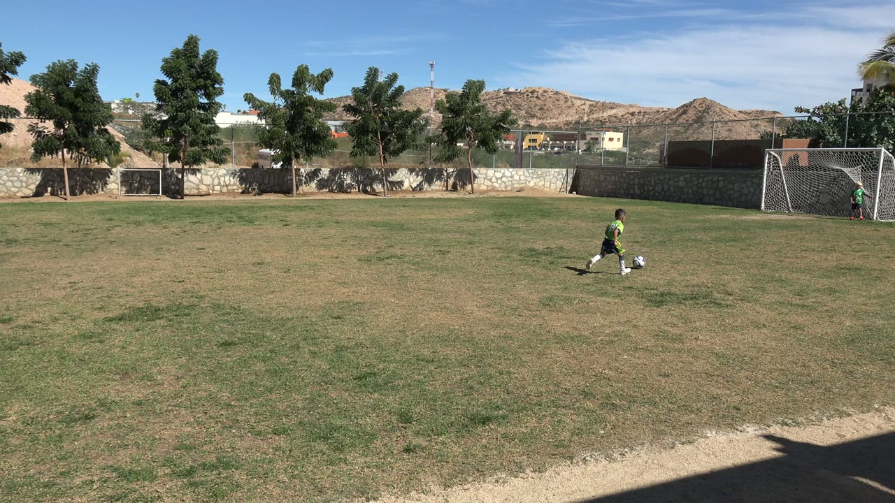 The boy dribbles the soccer ball across the sunlit Los Cabos field, with the open goal area and dry hills set behind the grass.