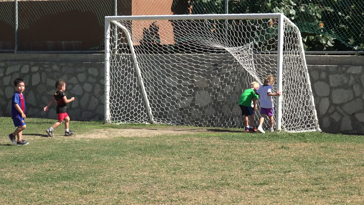 At 0:01 in Los Cabos, Mexico, two children stand tight to the right goalpost while another child crosses the grass in front of the net.