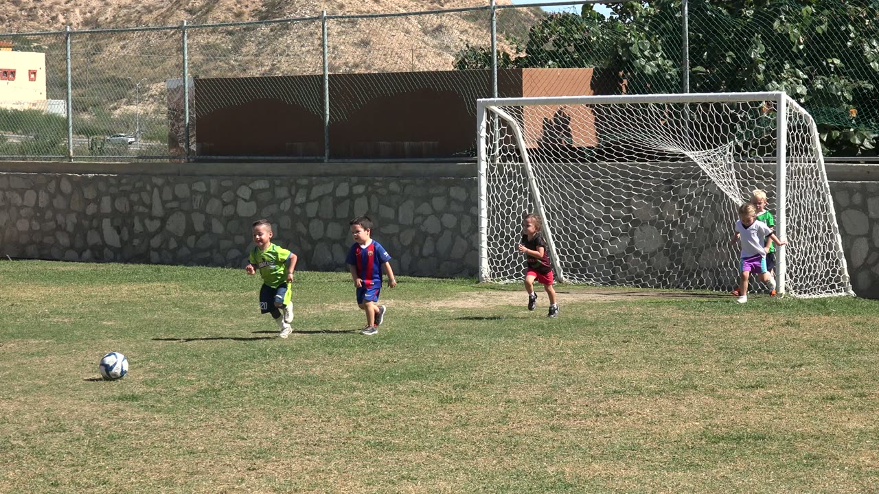 A child in a green uniform runs toward a soccer ball on a Los Cabos grass field while other children and the goal remain in the background.